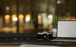 Modern dark hipster working desk with laptop white screen mockup, camera and empty space on tabletop over blurred night light background. 3d rendering, 3d illustration